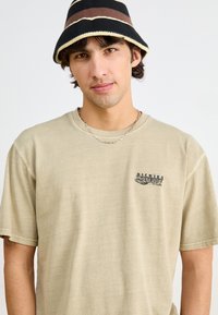 Tan short-sleeve t-shirt with a small black graphic logo. Worn with a black and brown striped bucket hat and layered silver chain necklace.