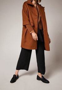 Abrigo de lana marrón con un cuello alto y bolsillos laterales, superpuesto a una blusa de color óxido y pantalones negros de pierna ancha. Zapatos loafers negros completan el conjunto.