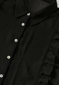 Camicia nera con colletto, realizzata in tessuto testurizzato, caratterizzata da pieghe verticali e cinque bottoni bianchi lungo la parte anteriore, con dettagli arricciati ai lati.