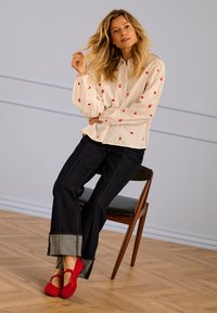 Vrouw met lang blond haar zittend op een houten stoel, gekleed in een witte blouse met rode hartjes, donkere spijkerbroek en rode Mary Janeschoenen.