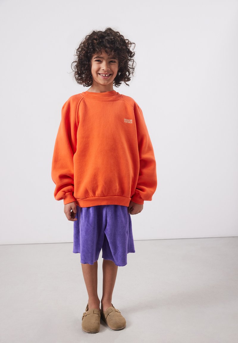 Dijete s kovrčavom kosom koje nosi preveliku narančastu trenirku, ljubičaste kratke hlače i bež cipele na navlačenje, stoji i smiješi se.