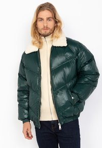 Green puffer jacket with a shiny finish, featuring a beige faux fur collar and ribbed cuffs. Worn over a cream zip-up sweater.