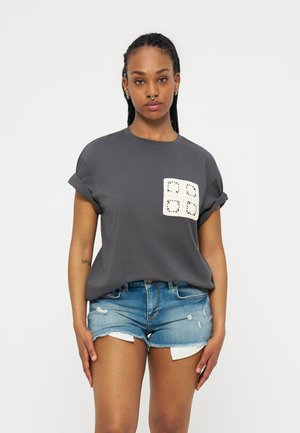 Kvinde med flettet hår iført en mørkegrå t-shirt med opkrullede ærmer, en hæklet firkantet lap og slidte denimshorts.