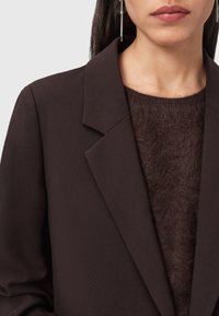 Blazer marrone aderente con rever a punta, indossato sopra un maglione scuro marrone a trama. Capelli neri lunghi e lisci e orecchini pendenti visibili.
