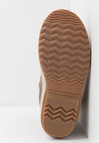 Suela de goma marrón con un patrón de zigzag, con el nombre de la marca "SOREL" grabado en la parte trasera. La suela está inclinada sobre un fondo blanco.