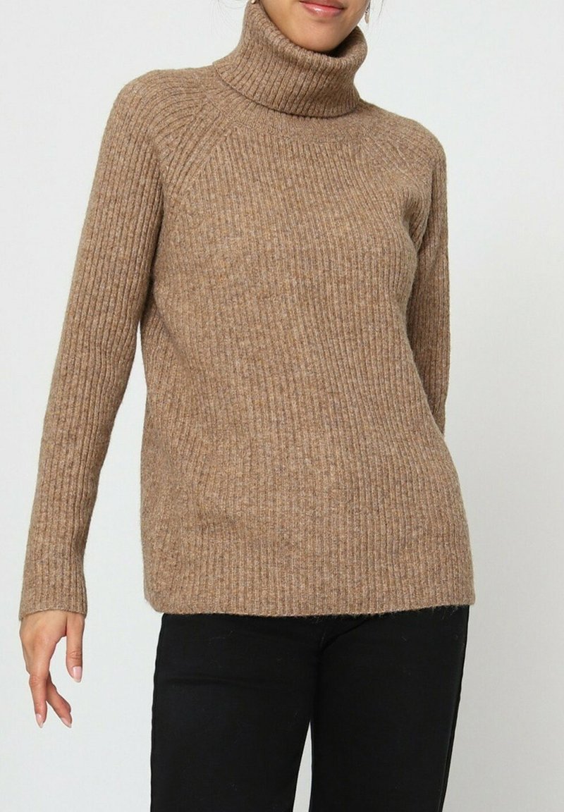 Femme portant un pull col roulé camel à côtes, associé à un pantalon noir, sur un fond clair et uni.