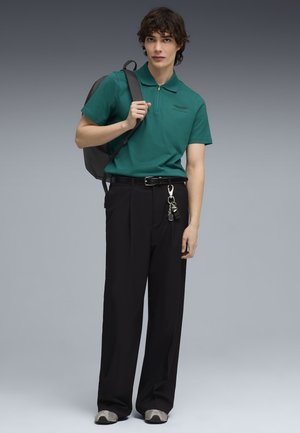 Jeune homme portant un polo vert, un pantalon noir avec un porte-clés, des baskets grises et un sac à dos noir porté sur une épaule.