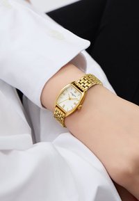 Goldfarbene Armbanduhr mit rechteckigem Zifferblatt, weißem Ziffernblatt und römischen Ziffern. Verfügt über ein Gliederarmband aus Metall und eine Krone.