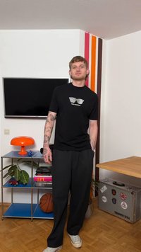 Νεαρός άνδρας με τατουάζ φορά μαύρο πουκάμισο και παντελόνι Karl Lagerfeld, στέκεται σε μοντέρνο δωμάτιο με πορτοκαλί λάμπα, μπάλα του μπάσκετ και ριγέ διακόσμηση στον τοίχο.