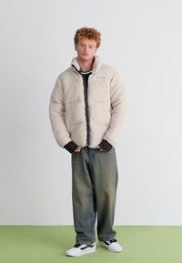 Puffer jacket in beige fleece with a front zipper, ribbed black cuffs, and gray lining. Paired with loose, faded green jeans and white sneakers.
