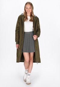 Olíva zöld hosszú kabát szürke kapucnival, cipz�árakkal és bordázott mandzsettával, szürke plisszírozott szoknyával és fehér sportcipőkkel bézs betétekkel párosítva.