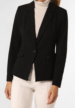 Femme portant un blazer noir ajusté boutonné une fois sur un col roulé blanc et un pantalon blanc, debout sur un fond uni.