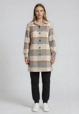 Donna che indossa un lungo cappotto beige, grigio e marrone a quadri con bottoni, pantaloni neri e scarpe da ginnastica bianche, in piedi davanti a uno sfondo semplice.