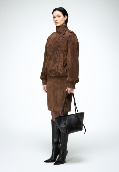 Vrouw in een bruine suède jas en rok, houdt een zwarte leren handtas vast, draagt zwarte kniehoge laarzen, staande tegen een effen achtergrond.