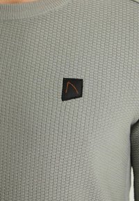 Grå stickad tröja med ribbad textur, rund halsringning och en svart fyrkantig lappar med en orange design detalj.