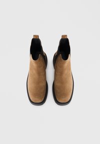 O pereche de ghete Chelsea din suede crem, cu panouri elastice laterale negre și talpă neagră, pe fundal alb, văzute din partea de sus.