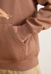 Sweat à capuche marron avec poche avant, présentant une texture lisse et un petit logo sur le poignet. Il a une coupe décontractée et un ourlet côtelé.