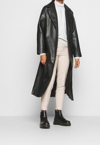 Cappotto lungo in ecopelle nera con ampi revers, indossato sopra un pullover grigio. Abbinato a pantaloni slim-fit beige chiaro e stivaletti neri.