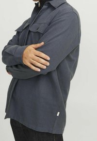 Grå skjorta med en texturerad tyg, två bröstfickor, krage med vikning och en något rundad fåll, bärs över mörka jeans.