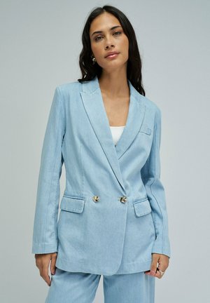Femme aux longs cheveux foncés portant un blazer croisé bleu clair et un pantalon assorti sur un fond uni.