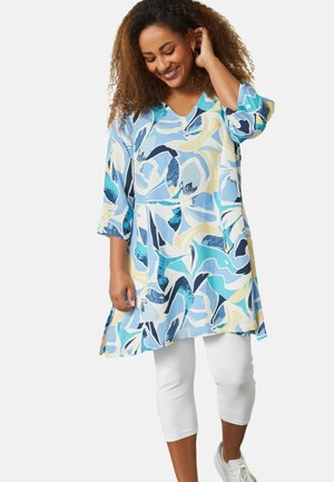 Blumige Tunika in Blau-, Gelb- und Grüntönen, mit V-Ausschnitt, Dreiviertelärmeln und A-Linie-Siluette, kombiniert mit weißen Leggings.