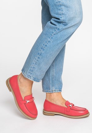 Person trägt hellblaue Jeans und leuchtend pinke Leder-Loafer mit beigefarbenen Sohlen, steht auf weißem Hintergrund.