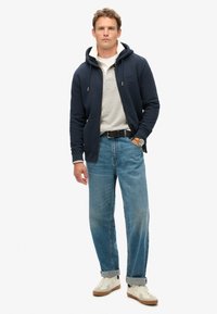 Granatowa bluza z kapturem na zamek z troczkami, jasnoszara koszulka polo pod spodem, jasnoniebieskie prostokątne dżinsy i kremowe sneakersy. Brązowy skórzany zegarek jako dodatek.