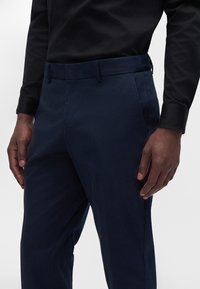 Homme portant un pantalon bleu marine foncé et une chemise noire à manches longues, debout avec les mains détendues sur les côtés.