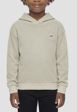 Dijete odjeveno u bež teksturiranu hoodicu s AM logom na prsima i crne hlače, stoji ispred neutralne pozadine.