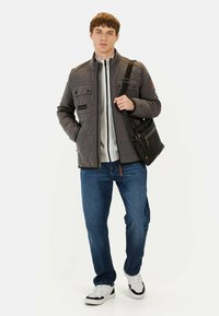 Grå quiltet jakke med frontlommer, hvid zip-up sweater, blå jeans og sort messenger taske. Modellen bærer hvide sneakers med sorte detaljer.