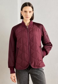 Giacca trapuntata bordeaux con zip frontale, polsini a costine e tasche laterali. Presenta un motivo testurizzato e una tasca con zip sulla manica.