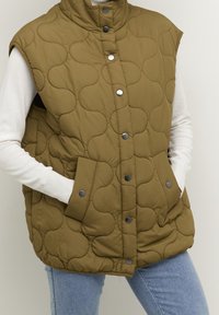 Chaleco acolchado sin mangas color verde oliva con cuello alto, botones a presión y dos bolsillos frontales, con un exterior suave y texturizado.