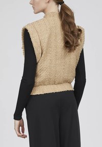 Beige tekstureret vest med et vævet stof, ærmeløs design, og høj krave, parret med en sort langærmet top under.