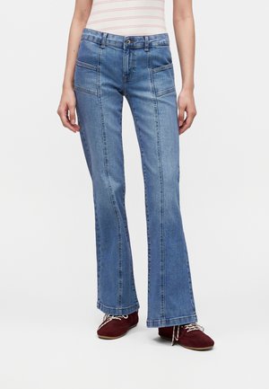 Femme portant un jean flare taille haute bleu clair avec poches plaquées devant et des chaussures rouges à lacets, debout devant un fond clair uni.