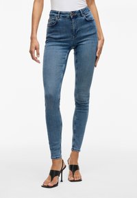 Hoge taille blauwe jeans van denimstof met een slanke pasvorm, voorzien van voorkeuzepockets en een knoopsluiting, gecombineerd met zwarte hoge hakken.