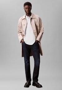 Light beige trench coat over a white polo shirt, paired with dark blue jeans and black shoes. The coat features a double-breasted design.