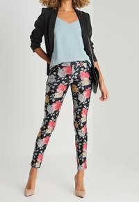 Blumenhose mit schwarzem Hintergrund, die rosa, graue und goldene Blumen zeigt. Kombiniert mit einem hellblauen Oberteil und einem schwarzen Blazer.