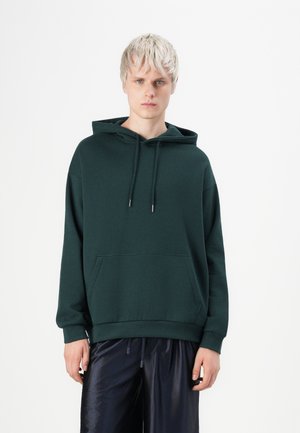 Νεαρό άτομο με κοντά ξανθά μαλλιά που φοράει σκούρο πράσινο φούτερ με κουκούλα και μαύρο γυαλιστερό παντελόνι, στέκεται μπροστά σε απλό λευκό φόντο.