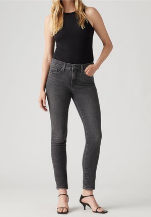 Femme portant un haut noir sans manches, un jean skinny gris foncé et des sandales noires à talons et brides, debout avec une main dans la poche.