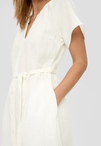 Witte, knoopsluiting jurk van textuurstof. Heeft korte mouwen, een ceintuur in de taille en zijzakken. Eenvoudig, casual ontwerp.