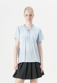 Blusa azzurro chiaro a maniche corte con scollatura arricciata, dettaglio a chiave e tessuto testurizzato. Abbinata a una gonna plissettata scura.