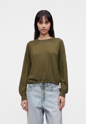Femme portant un pull vert olive à manches longues avec une taille à cordon et un jean taille haute bleu clair, debout devant un fond uni.