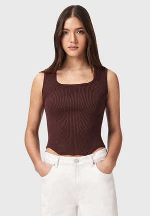 Jonge vrouw met lang bruin haar, gekleed in een mouwloze, bruin geribbelde top en witte broek, met handen in de zakken.