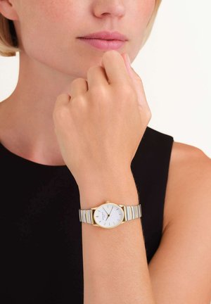 Montre en métal argenté et doré avec un cadran blanc, des index horaires minimalistes et un boîtier rond, portée au poignet d'une personne.