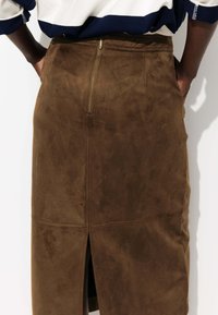 Jupe crayon en suede marron avec fermeture éclair au dos et fente. Texture lisse avec coutures visibles, épousant étroitement le corps.