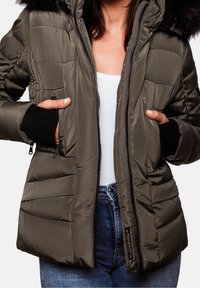 Vrouw in een opengezipte olijfgroene pufferjack met een zwarte bontkap, zwarte gebreide manchetten, een witte shirt en blauwe jeans, met handen in de zakken van de jas.