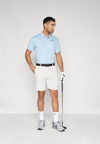 Camisa polo azul claro com um logotipo preto, combinada com calções brancos. Ele usa sapatilhas cinzentas e segura um taco de golfe, com um cinto preto.