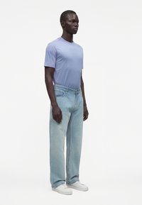 Hombre de pie con camisa de manga corta azul claro, vaqueros de pierna ancha de lavado claro y zapatillas blancas sobre un fondo blanco liso.