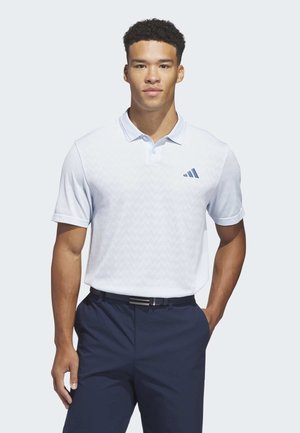 Uomo che indossa una polo bianca a maniche corte con un piccolo logo e pantaloni blu navy, in piedi con una mano in tasca.