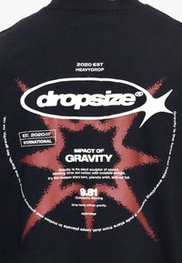 T-shirt noir avec texte graphique blanc et rouge sur la gravité, avec une explosion d'étoile rouge derrière le mot « dropsize » à l'intérieur d'une forme ovale.